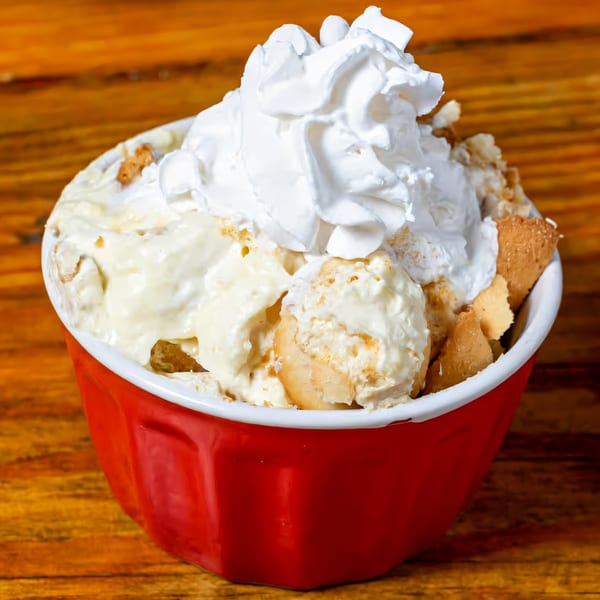 Banana pudding with whipped cream and vanilla wafers in a red bowl on a wooden table.